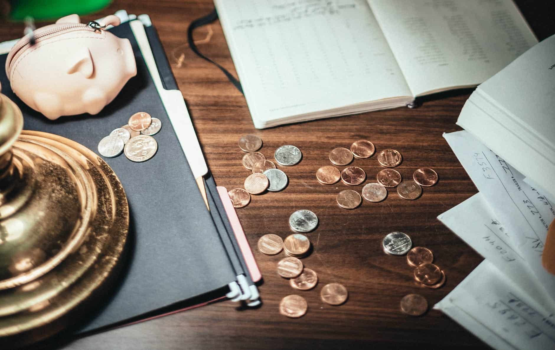 a brown desk which has got coins spread over it, with a piggy bank and notepads spread over the table.