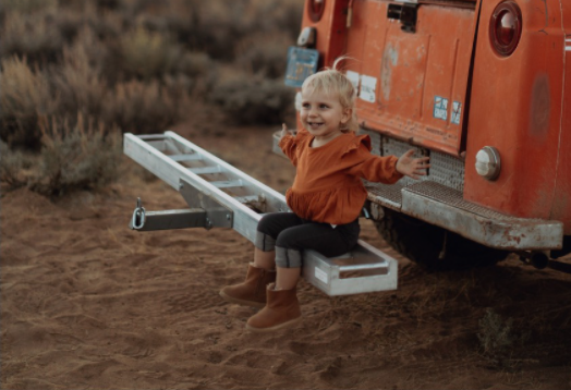 little girl sitting on the back bumper of a car, hands outstretched looking happy