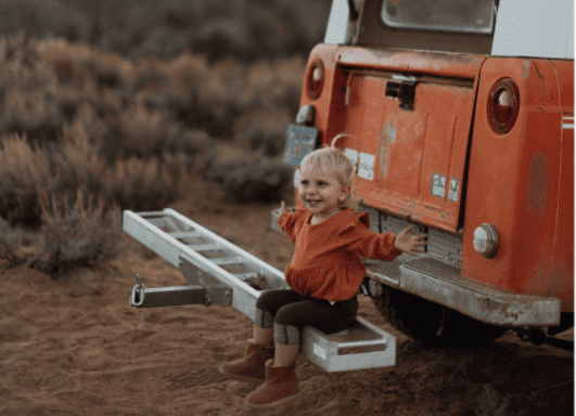 little girl sitting on the back bumper of a car, hands outstretched looking happy
