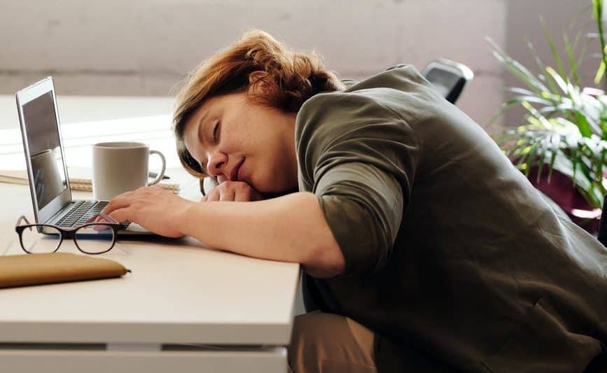 a lady asleep at her desk and her head is resting on her arm. Laptop is open in front of her with a cup