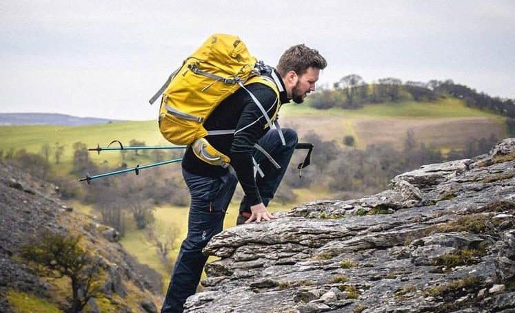 A man with hiking sticks climbing up a mountain with a yellow rucksack on his back.