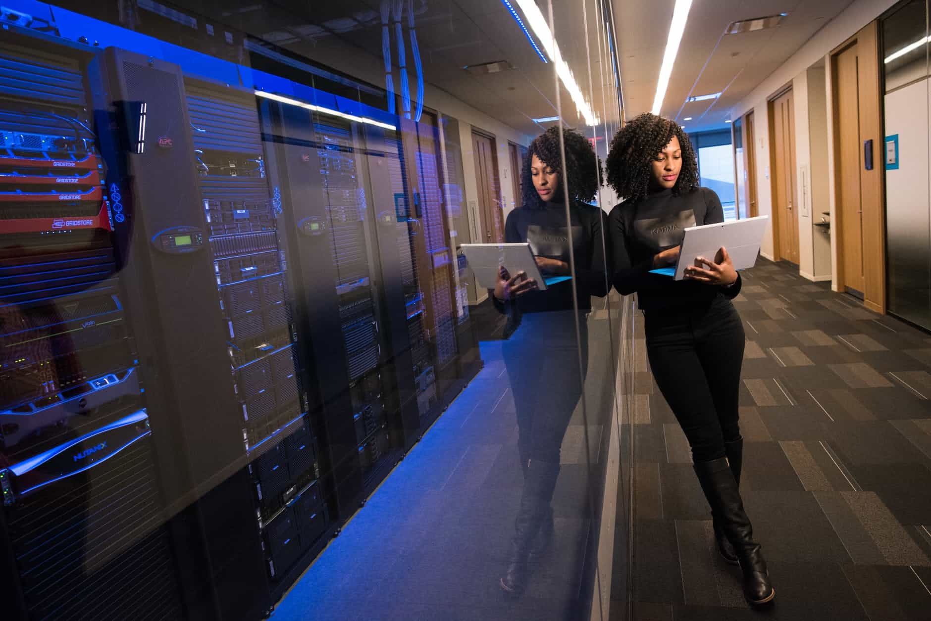 A woman leaning on a glass wall in the hallway of an office building while checking her laptop.