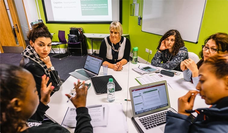 6 women sitting around a table, with laptops open, discussing meeting