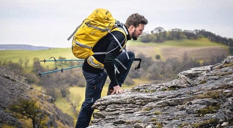 man with hiking sticks climbing up a mountain with a yellow rucksack on his back