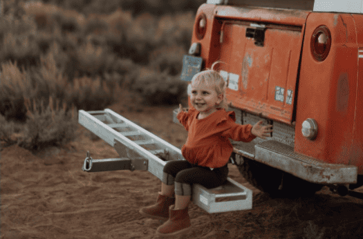 little girl sitting on the back bumper of a car, hands outstretched looking happy