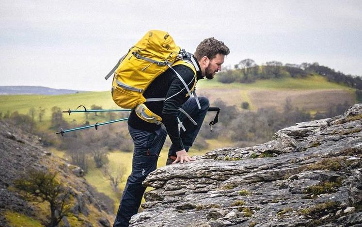 man with hiking sticks climbing up a mountain with a yellow rucksack on his back