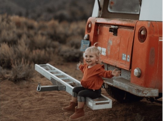 little girl sitting on the back bumper of a car, hands outstretched looking happy