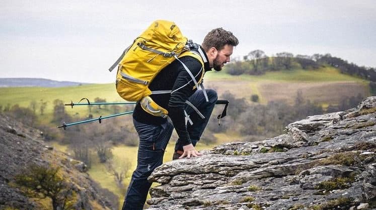 man with hiking sticks climbing up a mountain with a yellow rucksack on his back