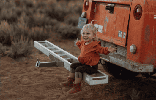 little girl sitting on the back bumper of a car, hands outstretched looking happy