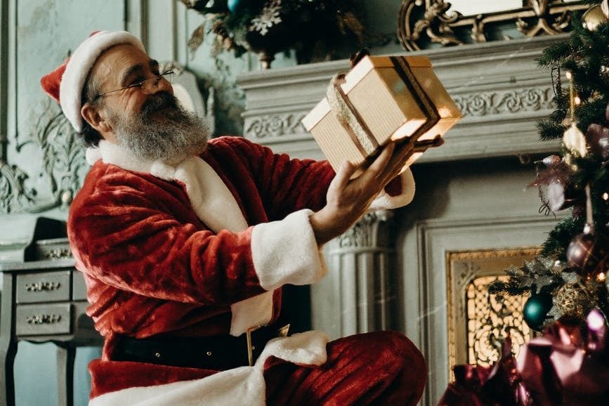 Father Christmas holding a present up to his face, whilst in a living room surrounded by Christmas decorations and a Christmas tree