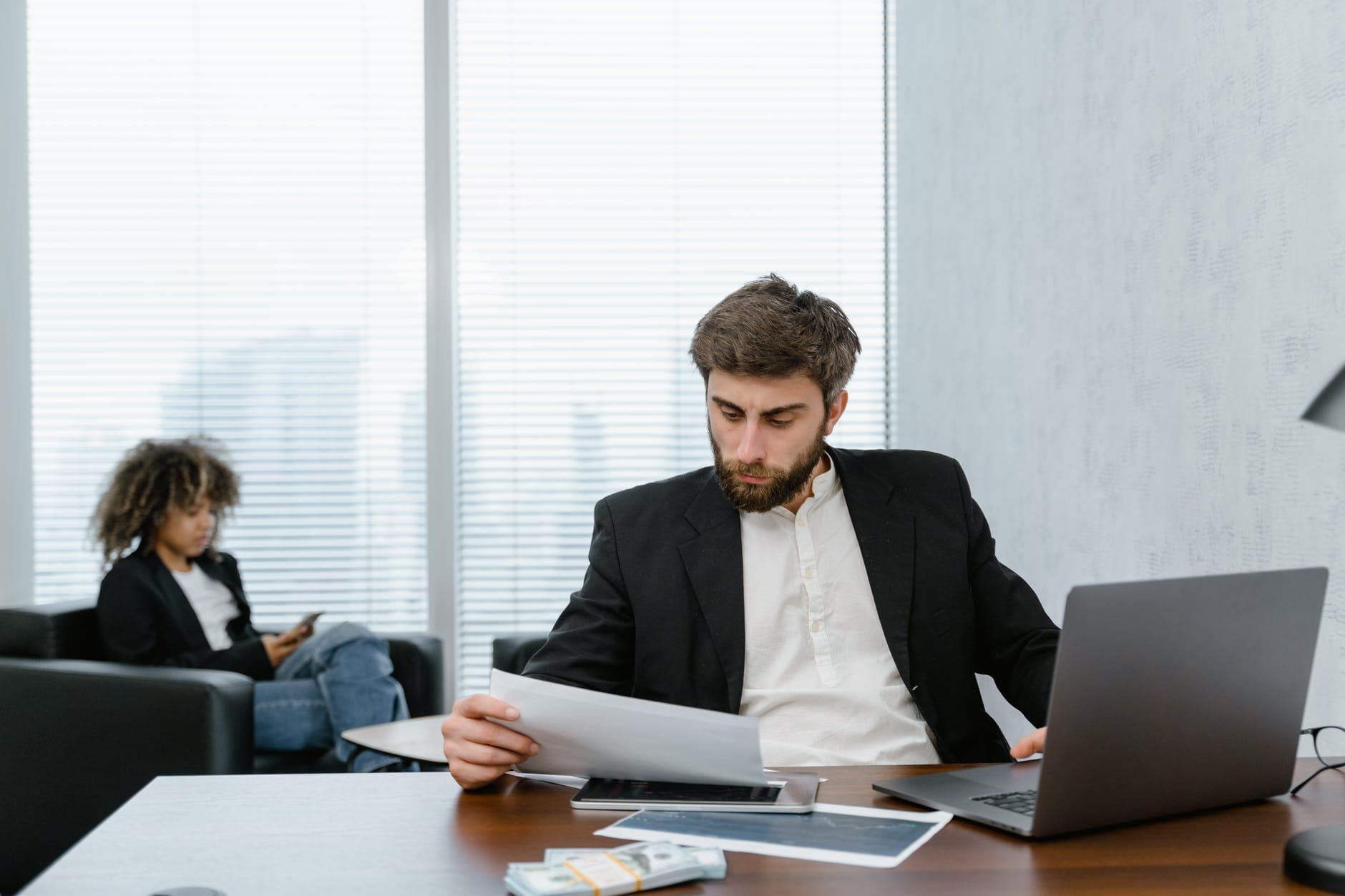 man sat at a desk looking at a piece of paper, with laptop open next to him. Woman in the background sat in a chair in an office.