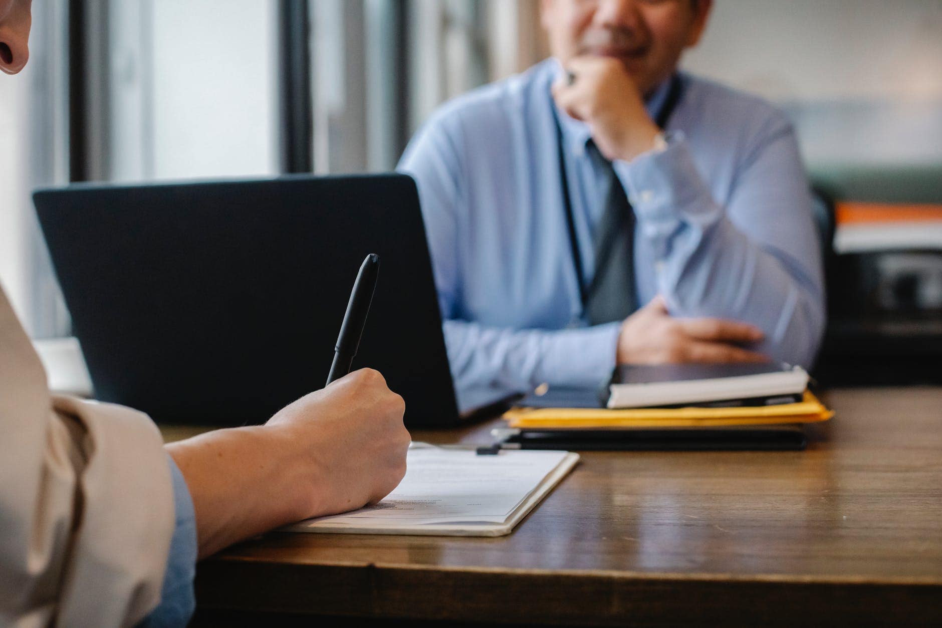 2 people at a desk, one is writing in a notebook with a pen in her right hand, the other person looks to be talking to her, with his laptop open in front of him