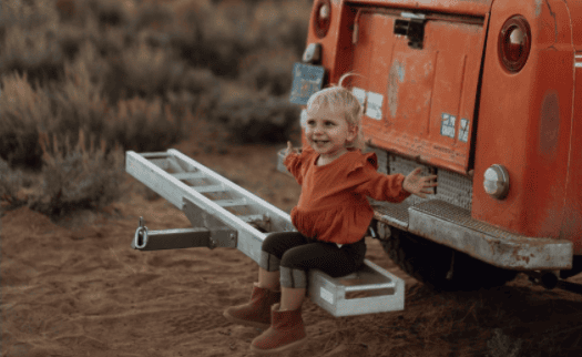 little girl sitting on the back bumper of a car, hands outstretched looking happy