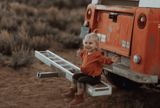 little girl sitting on the back bumper of a car, hands outstretched looking happy