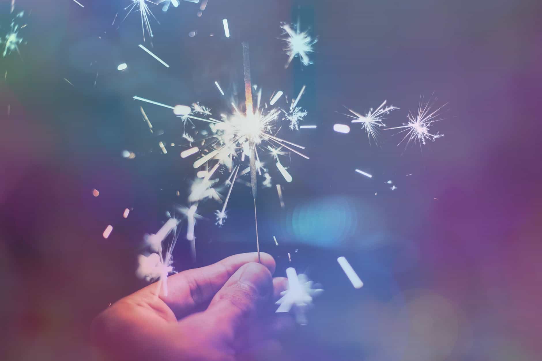 A hand holding a lit sparkler that is sparking brightly in the dark.