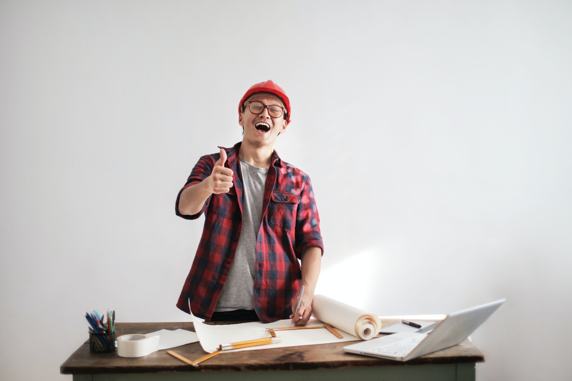 A man standing and working on building plans at a desk, he is wearing a construction helmet, smiling and giving a thumbs up.