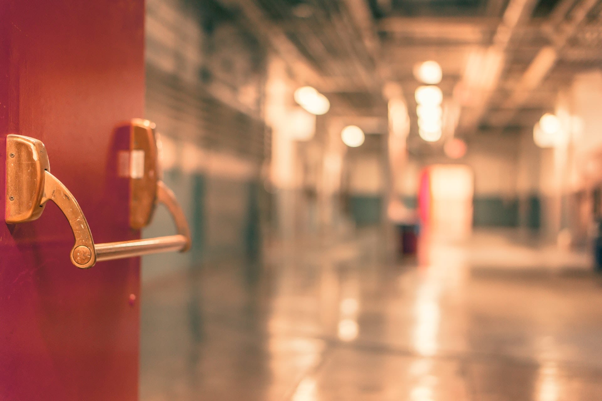 An out-of-focus corridor with a red push bar door on the left.