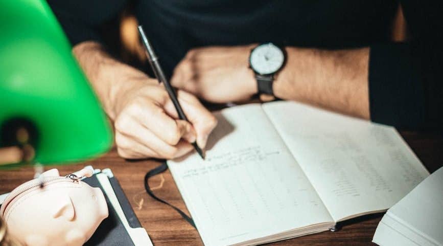 A man writing in notebook at his desk. There is a lamp and piggy bank on the desk.