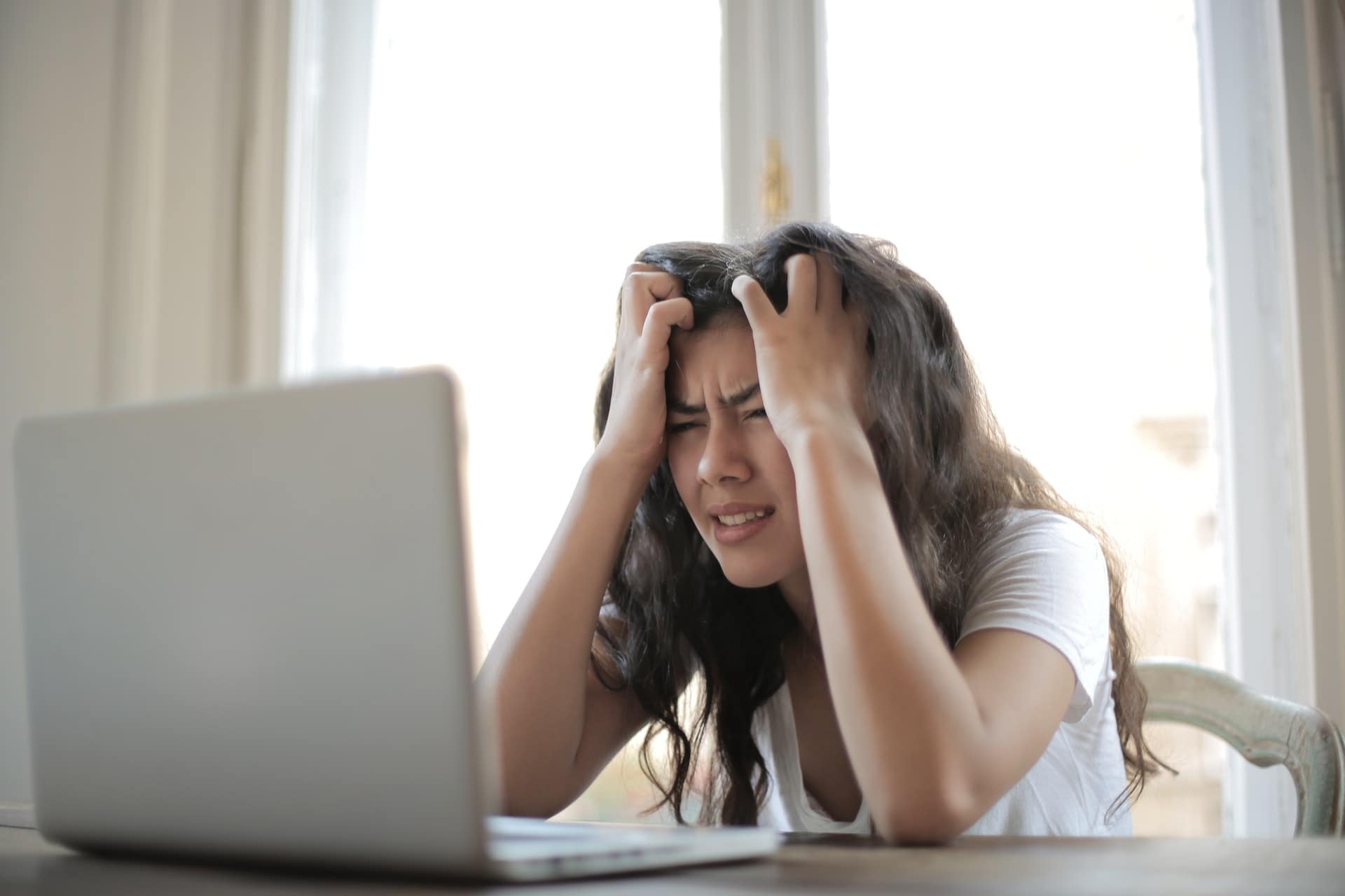 A woman looking at her laptop with a confused look on her face while holding her head in her hands, she is sitting at a desk.