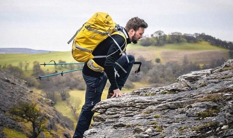 man with hiking sticks climbing up a mountain with a yellow rucksack on his back