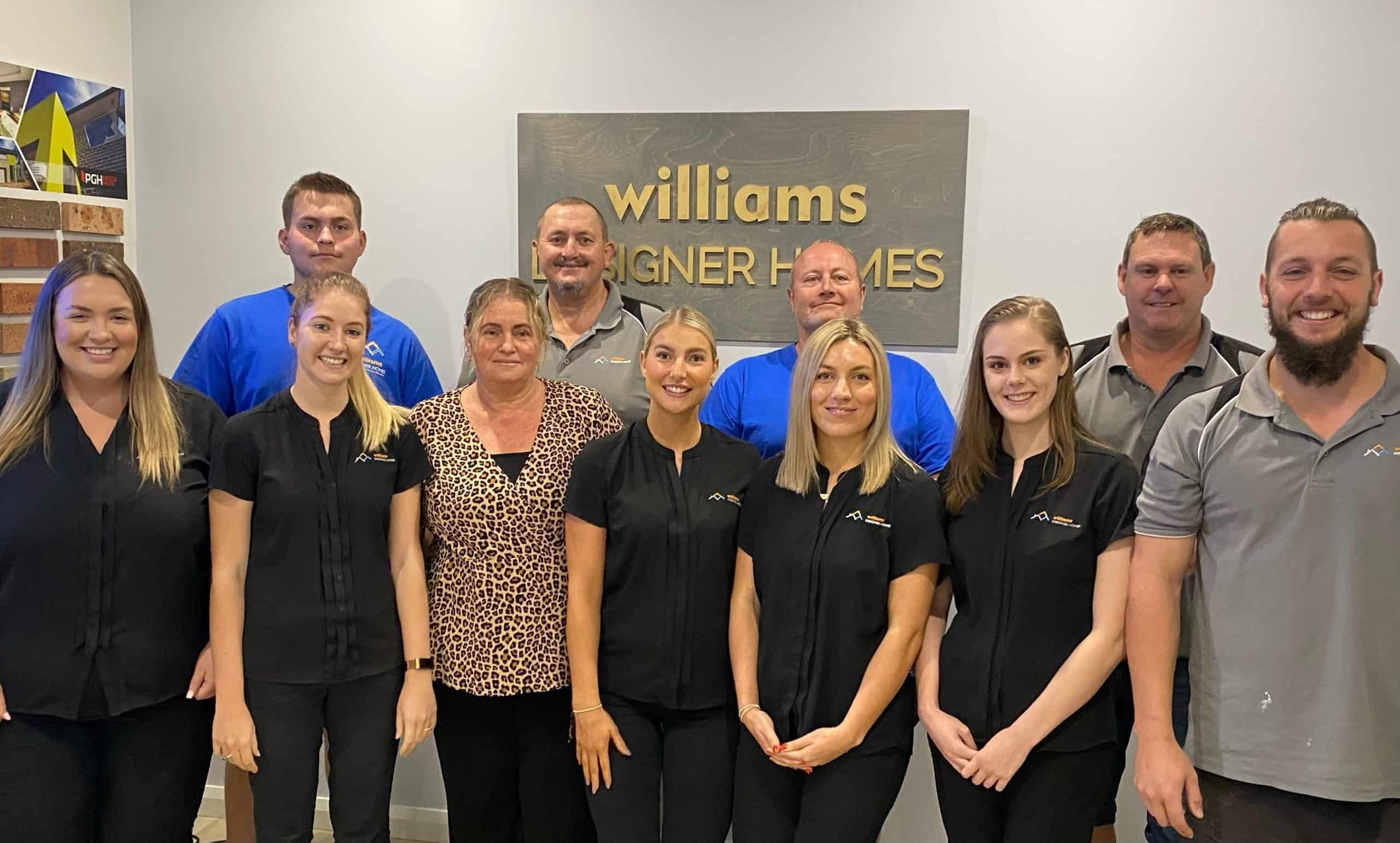 William Designer Homes team standing in front of their office wall with a plaque of their behind them.