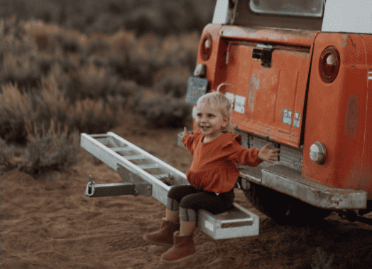 little girl sitting on the back bumper of a car, hands outstretched looking happy